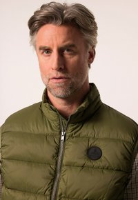 Olivově zelená vycpaná vesta s předním zipem, vysokým límcem a kulatým černým logem na hrudi. Texturovaná tkanina s kostkovanou podšívkou.