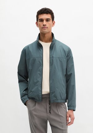 Mann trägt eine türkisfarbene Zip-up-Jacke über weißem Hemd und grauer Hose, steht mit einer Hand in der Tasche vor einfachem hellem Hintergrund.