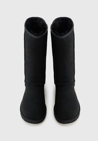 Svarta knähöga stövlar med en slät yta i mocka, mjukt foder av konstpäls, rundad tå och sydda detaljer längs sömmarna.