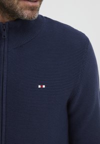 Mornarsko modra zip-up jopica z rebrasto teksturo, visokim ovratnikom in majhnim logotipom z rdečimi, belimi in modrimi akcenti na prsih.