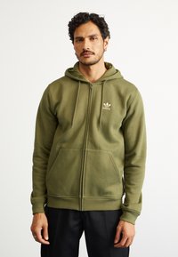 Corta-vento verde-azeitona com fecho zip em mistura de algodão, apresentando um bolso frontal, cordões e um pequeno logo branco no peito. Textura suave.