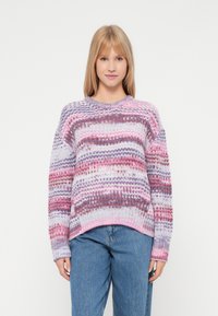 Young woman with straight blonde hair wearing a colorful striped knit sweater and blue jeans, standing against a plain white background.