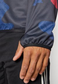 Giacca sportiva nera con un tessuto grigio testurizzato caratterizzato da motivi astratti blu e rossi; polsini elastici con dettagli neri visibili.