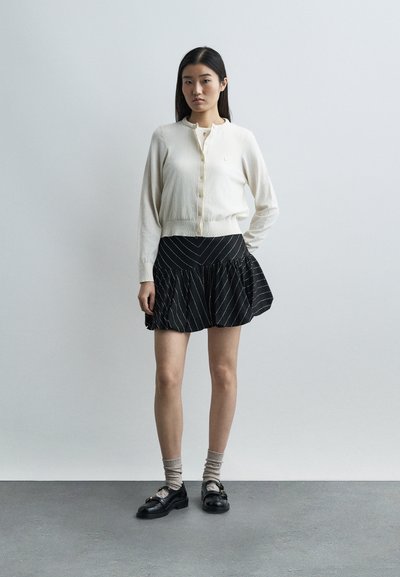 Νέα γυναίκα που φοράει λευκή ζακέτα, φούστα με μαύρες ρίγες, μπεζ κάλτσες και μαύρα λοφέρ, στέκεται μπροστά σε έναν απλό φωτεινό τοίχο.