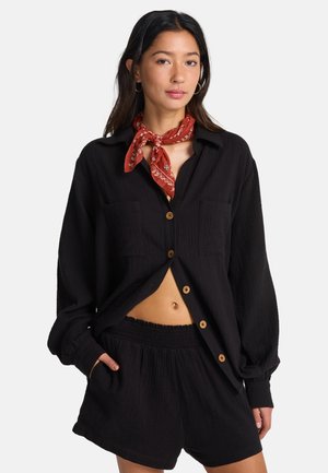 Camicia nera leggera con bottoni, maniche lunghe e due tasche sul petto, abbinata a pantaloncini neri coordinati con vita elastica. Un fazzoletto rosso completa l'outfit.