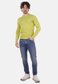 Uomo con capelli ricci che indossa un maglione verde lime, jeans blu e sneakers bianche, in piedi con una mano in tasca su uno sfondo bianco.