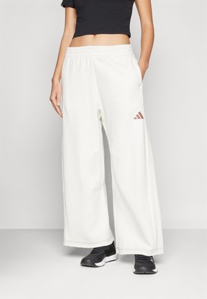 Femme portant un pantalon blanc à jambes larges avec une taille élastique et un petit logo sur la cuisse, associé à un crop top noir et des baskets noires.