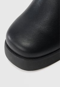 Melns ādas potītes zābaks ar gludu virsmu, noapaļotu purngalu un izturīgu gumijas zolīti. Raksturo minimālistisks dizains un bezšuvju malas.