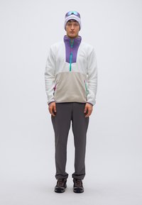 Ελαφρύ φλις μπλούζα σε λευκό χρώμα με μοβ πανελ και πράσινο φερμουάρ, σεταρισμένη με γκρι παντελόνι και μαύρα ορειβατικά παπούτσια. Αθλητικά γυαλιά ηλίου και σκουφί.