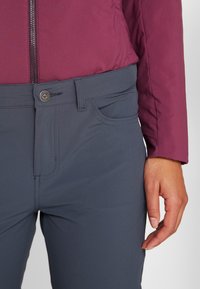 Mörkgrå byxor med en slät textur, med framknäppning och sidofickor, parat med en vinröd zip-up jacka.