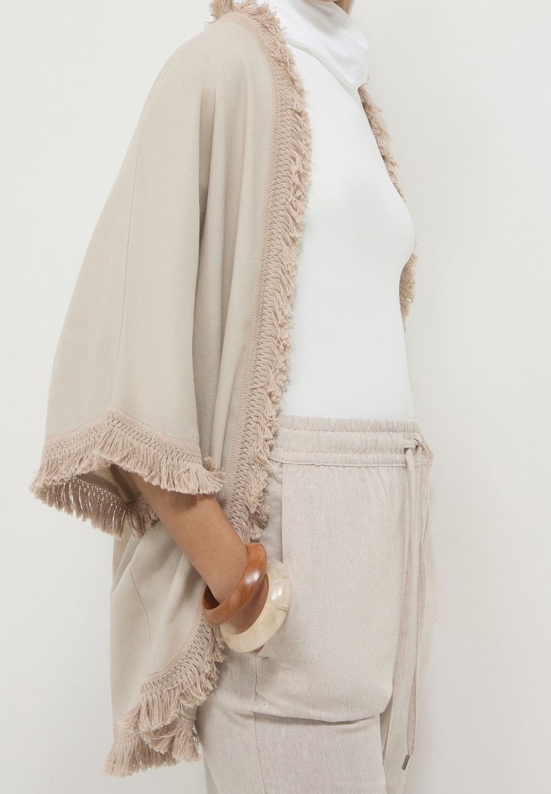 Beige kimono-stijl omslag met franjes, gedragen over een witte top. Gecombineerd met textuur, lichtgekleurde broeken en gestapelde houten armbanden.