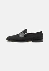 Mocasín de tela negra con una forma elegante, que presenta un acento negro brillante sobre el empeine y una suela de goma plana.