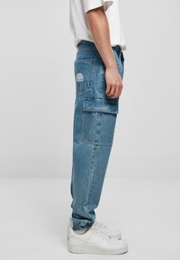 Lysblå denim cargo bukser med store sidelommer, elastiske manchetter, og hvid broderet logo detalje. Sættes sammen med hvide sneakers.