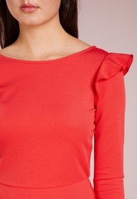 Haut rouge à manches longues avec un col rond et des détails de manches froncées. Le tissu semble lisse avec une silhouette ajustée.