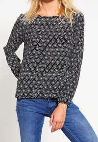 Svart långärmad blus med ett vitt stjärnblossmönster, rund halsringning och elastiska ärmslut, i kombination med blå jeans.