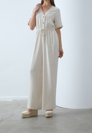 Femme portant une combinaison en lin beige avec des boutons et une taille à cordon, associée à des chaussures à talon compensé en jute beige, se tenant sur un sol en béton.