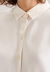 Close-up of a person wearing a cream-colored button-up shirt with a pointed collar and visible buttons down the front.