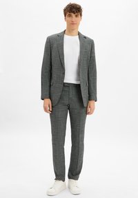 Junger Mann in einem grauen karierten Anzug mit einem weißen T-Shirt und weißen Sneakers, der vor einem schlichten weißen Hintergrund steht.