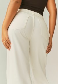 Valkoiset leveälahkeiset housut, joissa on korkea vyötärö, kaksi takataskua ja sileä, teksturoitu kangas. Säädettävä vyötärönauha on näkyvissä.