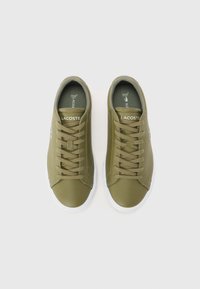 Sapatilhas verde-azeitona com parte superior de material sintético suave, sola de borracha branca e fecho com atacadores. Apresentam um logótipo da Lacoste na lateral e colarinho acolchoado.