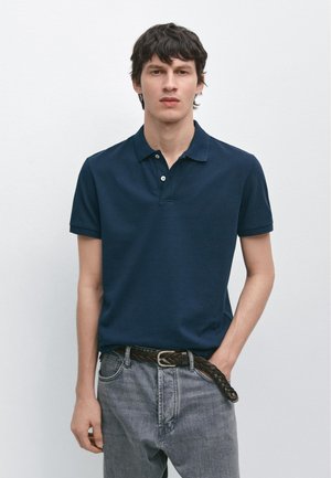 Giovane uomo con indosso una polo blu navy a maniche corte infilata in un paio di jeans grigi sbiaditi con una cintura nera intrecciata, in piedi davanti a uno sfondo semplice.