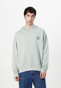 Hellgrüner Hoodie aus weichem Stoff, mit lockerer Passform, Kapuze mit Kordelzug und einem schwarzen Druck auf der linken Brust.
