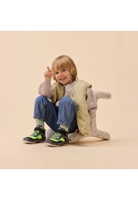 Ein Kind trägt eine hellgrüne gesteppte Weste über einem grauen Sweatshirt, blauen Jeans und schwarzen Schuhen mit grünen Akzenten und sitzt auf einem bunten, strukturierten Hocker.