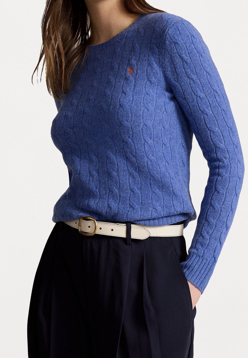 Femme portant un pull torsadé bleu ajusté avec un petit logo rouge, rentré dans un pantalon noir taille haute avec une ceinture beige.