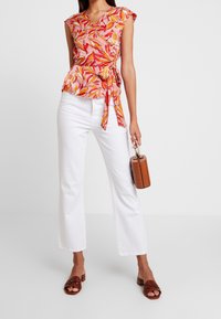 Blomstret kortærmet top i pink, orange og gul med peplum talje, parret med hvide flared bukser og brune slidesandaler. Bærer en lille brun taske.