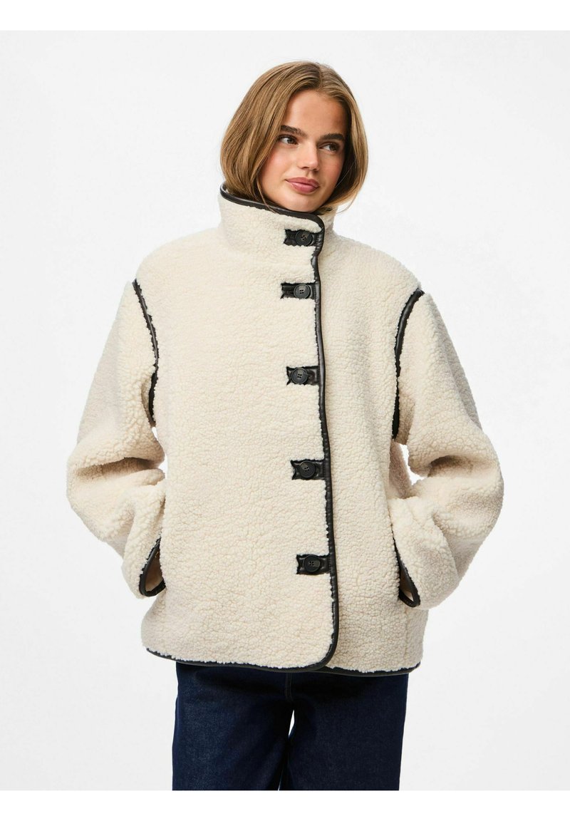 Kremfarget fleecejakke med høy krage, svarte knappedetaljer og kontrastsøm. Løs passform med to sidelommer.