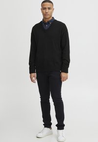 Pulover negru cu decolteu în V peste o cămașă albastră cu guler, asortat cu blugi închiși și adidași albi. Puloverul are manșete și tiv cu model rib. 