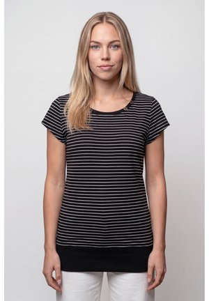 Blonde Frau, die ein schwarz-weiß gestreiftes Kurzarmshirt und weiße Hosen trägt, stehend vor einem einfarbigen hellen Hintergrund.