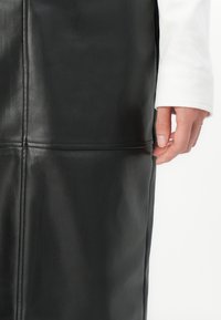 Sorte skinnskjørt med en jevn tekstur, strukturert design og synlige sømmer. Den ene hånden hviler på siden av skjørtet.