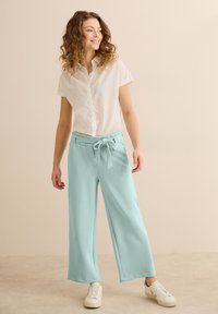 Chemise blanche légère à manches courtes associée à un pantalon large vert menthe à taille élastique avec un lien. Texture lisse et coupe décontractée.