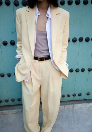 Adulto vistiendo un traje crema de gran tamaño con camisa blanca y camiseta gris, con las manos en los bolsillos, de pie frente a una puerta verde azulado con tachuelas negras redondas.