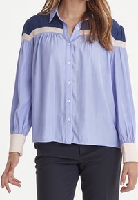 Jasnoniebieska koszula zapinana na guziki z granatowymi i beżowymi akcentami na ramionach, z długimi, bufiastymi rękawami i luźnym krojem. Gładka tekstura materiału.