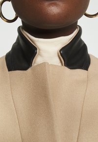 Gros plan sur une personne portant un manteau beige avec un col en cuir noir, un col roulé crème et des boucles d'oreilles anneaux dorés sur un fond blanc.