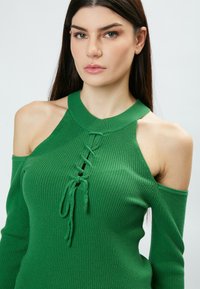 Grün gerippter Stricktop mit schulterfreiem Design, hoher Halslinie und einem vorn geschnürten Detail mit langen Bändern. Glatte Textur.