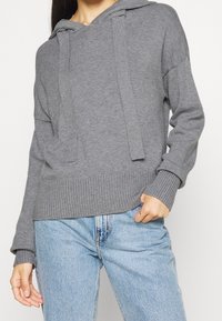 Grå huvtröja med ribbad stickad struktur, långa ärmar och snörningsdetalj. Matchad med ljusblå denimjeans.