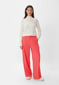 White knit turtleneck sweater paired with coral wide-leg trousers. Model wears light-colored shoes; the ensemble features smooth textures.