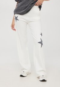 Białe dresowe spodnie z szarymi wzorami gwiazdek po bokach, wykonane z miękkiego materiału. W zestawieniu z ciemnoszarym, oversize'owym t-shirtem i białymi trampkami.