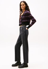 Cárdigan corto negro con patrones morados y rojos, combinado con jeans negros de cintura alta que presentan tachuelas a lo largo del lateral. Mocasines negros completan el conjunto.