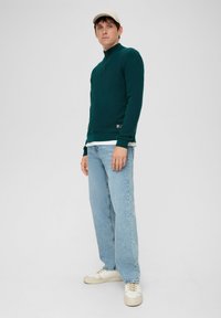 Dunkelgrüner Reißverschluss-Pullover aus strukturiertem Stoff, über einem weißen Hemd getragen, kombiniert mit hellblauen Jeans und cremfarbenen Sneakers.