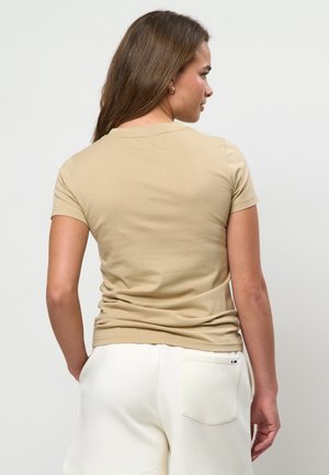 Kobieta o długich brązowych włosach, ubrana w beżową koszulkę z krótkim rękawem i białe szorty, stojąca tyłem na tle jasnego, jednolitego tła.