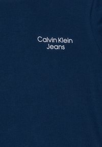 Marineblått tyg med mjuk textur som har en vit broderad "Calvin Klein Jeans"-logotyp. Enkel rund halsringning, minimalistisk design.