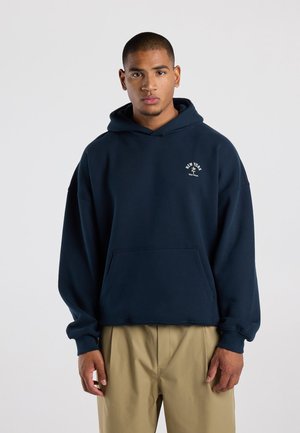 Junger Mann trägt einen dunkelblauen Hoodie mit kleinem weißem New York-Logo auf der Brust und beigen Hosen und steht vor einem einfarbigen Hintergrund.