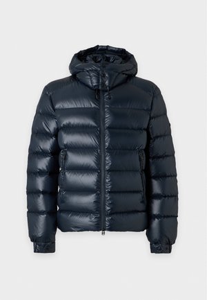 Homme portant une veste matelassée foncée, un polo marron, un pantalon beige et des mocassins marron foncé, debout devant un fond uni.