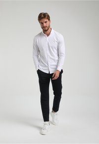 Camisa blanca de manga larga con botones, combinada con pantalones oscuros y zapatillas blancas. El conjunto es casual pero tiene un aspecto pulido.