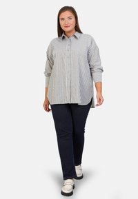 Chemise à rayures gris clair et blanche, à manches longues et à coupe décontractée, associée à un pantalon bleu foncé et des chaussures blanches.
