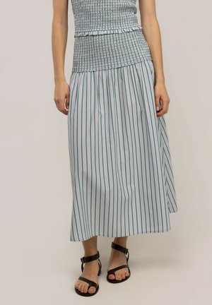 Mujer con una falda midi de rayas verdes y blancas, un top fruncido y sandalias negras con pulsera en el tobillo sobre un fondo neutro.